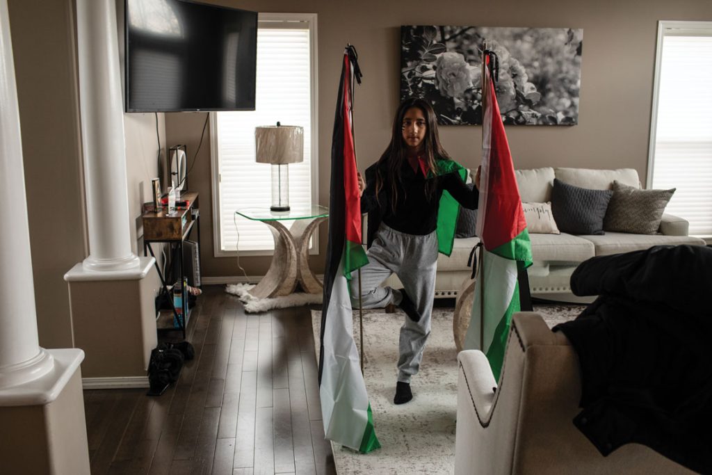 Zeher Assaf balancing on on leg in her living room. She is holding a Palestinian flag on either side to support her